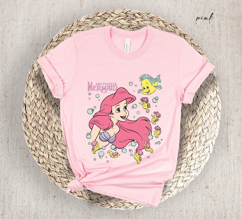 Peut inclure: T-shirt rose avec un motif d'Ariel, la Petite Sir&egrave;ne, avec Polochon et des hippocampes. Le texte "The Little Mermaid" est imprim&eacute; sur le t-shirt.