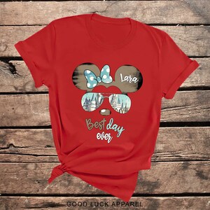 May include: Red t-shirt with a cartoon mouse head wearing sunglasses and a bow. The sunglasses have a castle image inside. The text "Best day ever" and "Lara" are on the shirt.