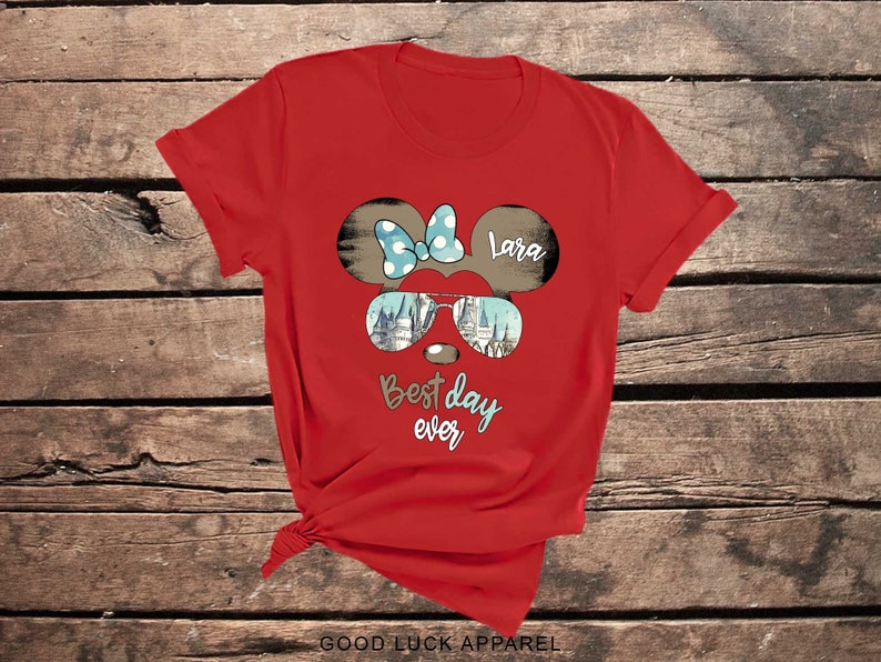 May include: Red t-shirt with a cartoon mouse head wearing sunglasses and a bow. The sunglasses have a castle image inside. The text "Best day ever" and "Lara" are on the shirt.