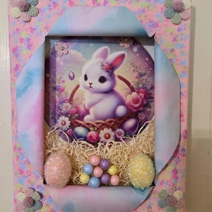 May include: A pink and blue framed picture with a white bunny rabbit in a basket of flowers. The picture is surrounded by pastel colored Easter eggs and a nest of straw.