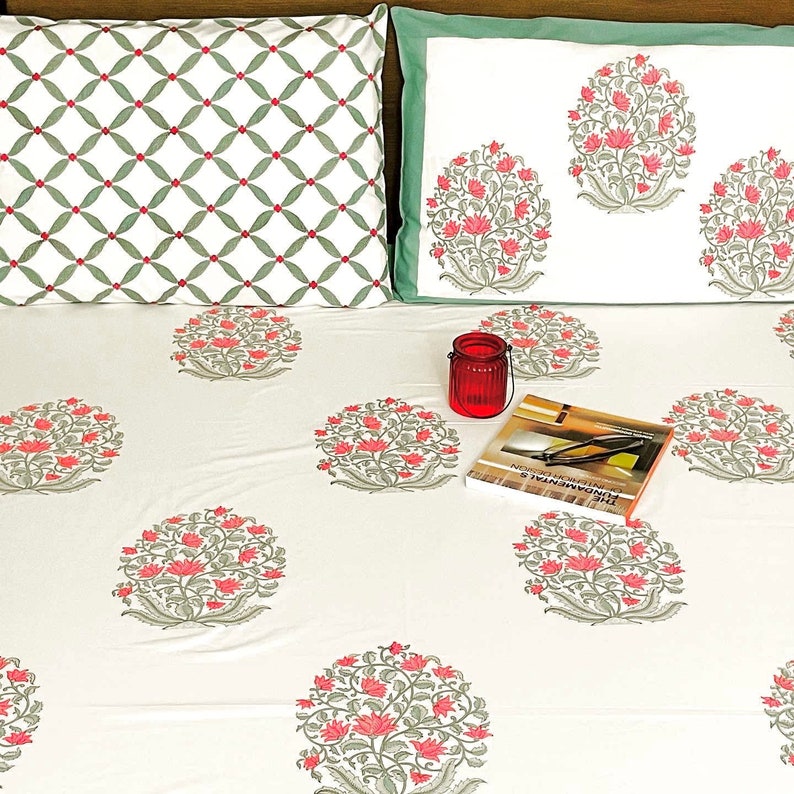 K&ouml;nnte beinhalten: Wei&szlig;e Bettw&auml;sche mit einem rosa-grauen Blumenmuster. Zwei Kissenbez&uuml;ge sind sichtbar, einer mit passendem Blumenmuster und der andere mit einem gr&uuml;n-wei&szlig;en geometrischen Muster. Ein roter Kerzenhalter und ein Buch liegen auf dem Bett.