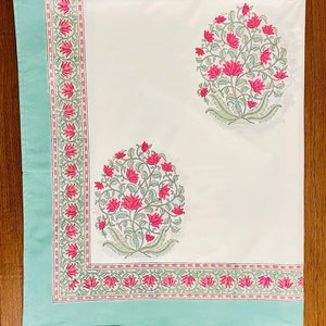 K&ouml;nnte beinhalten: Eine wei&szlig;e Tischdecke mit hellt&uuml;rkisfarbenem Rand und floralem Design. Der Rand weist ein sich wiederholendes Muster aus rosa Bl&uuml;ten und gr&uuml;nen Bl&auml;ttern auf. Zwei Gruppen aus rosa Bl&uuml;ten und gr&uuml;nen Bl&auml;ttern sind auf die Tischdecke gedruckt.
