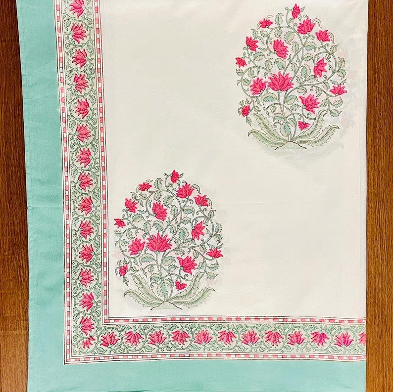 K&ouml;nnte beinhalten: Eine wei&szlig;e Tischdecke mit hellt&uuml;rkisfarbenem Rand und floralem Design. Der Rand weist ein sich wiederholendes Muster aus rosa Bl&uuml;ten und gr&uuml;nen Bl&auml;ttern auf. Zwei Gruppen aus rosa Bl&uuml;ten und gr&uuml;nen Bl&auml;ttern sind auf die Tischdecke gedruckt.