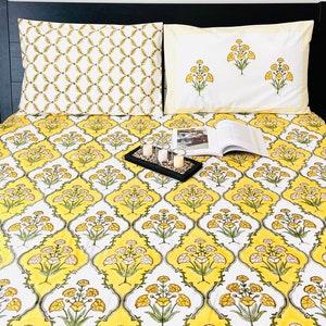 May include: A yellow and white floral patterned bedspread with a white pillow with yellow floral embroidery and a white pillow with a geometric pattern. There are four white candles on a black tray on the bedspread.