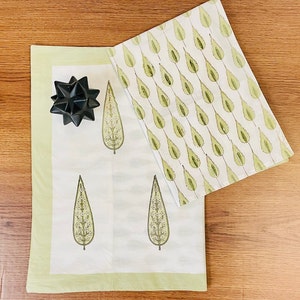 May include: Two white cotton tea towels with a green leaf pattern. One towel has a repeating pattern of leaves, while the other has three larger leaves with white flowers. The towels have a light green border.
