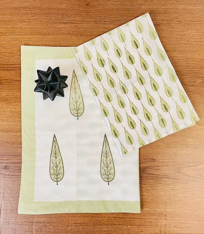 May include: Two white cotton tea towels with a green leaf pattern. One towel has a repeating pattern of leaves, while the other has three larger leaves with white flowers. The towels have a light green border.