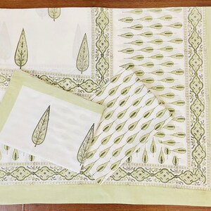 May include: A set of green and white cotton napkins with a leaf pattern. The napkins are folded and arranged on a green and white patterned fabric.
