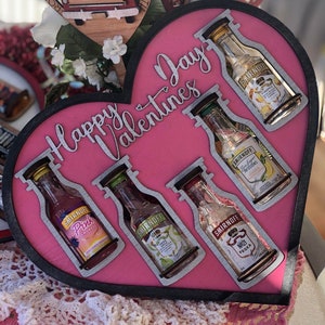 Może przedstawiać: Różowe, drewniane serce z napisem "Happy Valentine's Day" w srebrnych literach. Serce ma pięć wyciętych otworów na miniaturowe butelki alkoholu. Butelki to Smirnoff vodka, Smirnoff Pink Lemonade, Smirnoff Pineapple, Smirnoff No. 21 i Malibu rum.