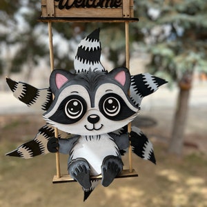 May include: A wooden raccoon figurine with a welcome sign above it. The raccoon is painted black, white, and gray and has large eyes. It is sitting on a swing.