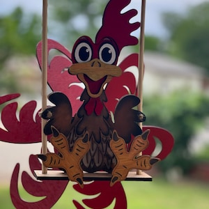 May include: A wooden rooster figurine with a red comb and wattle, brown feathers, and yellow feet, is swinging on a red and white wooden swing.