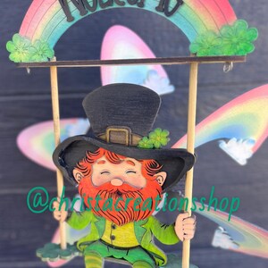 May include: A wooden leprechaun figurine on a swing with a rainbow arch above that says "WELCOME". The leprechaun is wearing a green hat, green pants, and a green vest. He has a red beard and a happy expression.