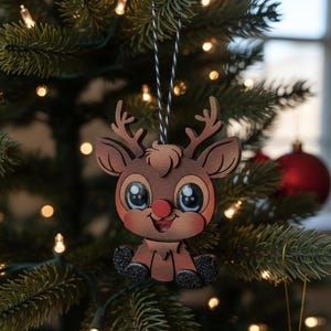 May include: A wooden Christmas ornament shaped like a cartoon reindeer with large blue eyes, a red nose, and glittery black feet. The ornament hangs from a black and white string on a Christmas tree with red ornaments and lights.