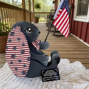 May include: A dark gray bunny figurine with an American flag design on one ear and a matching bow tie. The bunny holds a small American flag. The figurine sits on a white lace doily. The bunny is a patriotic decoration.