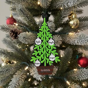 May include: A green Christmas tree ornament with a black outline, featuring white ornaments with names: Kyle, Kayle, Kooper, and Kason. The tree sits in a brown pot with the name Griffith. The ornament hangs on a decorated Christmas tree with red and gold ornaments.