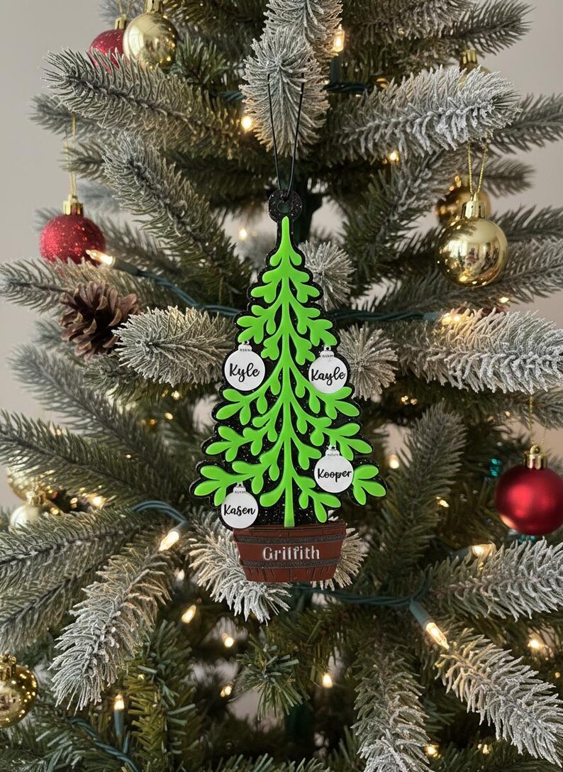 May include: A green Christmas tree ornament with a black outline, featuring white ornaments with names: Kyle, Kayle, Kooper, and Kason. The tree sits in a brown pot with the name Griffith. The ornament hangs on a decorated Christmas tree with red and gold ornaments.