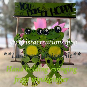 May include: Two green frogs holding hands on a swing, with a sign above them that reads "You make me hoppy". The frogs are sitting on a pink flower. The sign is made of wood and has a rustic look. The frogs are painted with black eyes and white pupils. The sign is painted with black letters on a green background. The frogs are holding hands and are smiling. The sign is hanging from a wooden stick. The frogs are a laser cut design file.