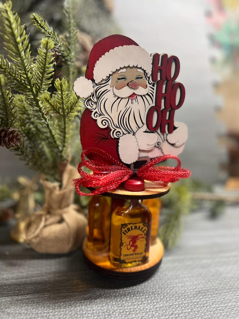 May include: Wooden Santa Claus figurine with a red hat and beard, holding a sign that says "Ho Ho Oh". The figurine is holding a bottle of Fireball Cinnamon Whisky. The figurine is on a wooden stand with a red ribbon.