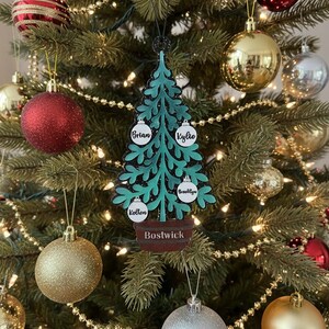 May include: A teal Christmas tree ornament with the names "Brian", "Kylie", "Brooklyn", and "Kotten" on white ornaments. The name "Bostwick" is on the base. The ornament hangs on a decorated Christmas tree with gold, silver, and red ornaments.