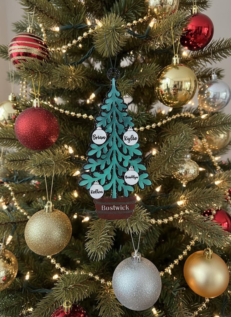 May include: A teal Christmas tree ornament with the names "Brian", "Kylie", "Brooklyn", and "Kotten" on white ornaments. The name "Bostwick" is on the base. The ornament hangs on a decorated Christmas tree with gold, silver, and red ornaments.