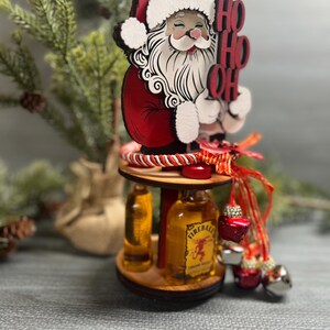 May include: A wooden Santa Claus figurine holding a sign that says "Ho Ho Oh" with two miniature liquor bottles on a wooden stand. The bottles are Fireball Cinnamon Whisky and a clear liquor bottle.