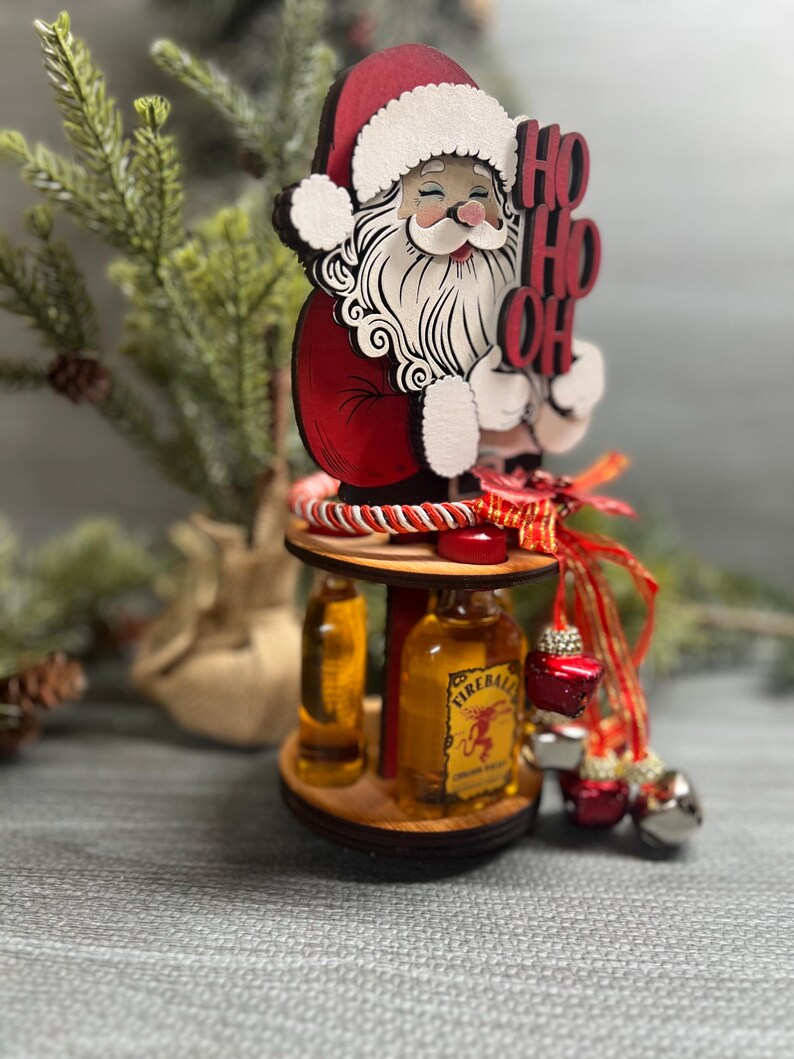 May include: A wooden Santa Claus figurine holding a sign that says "Ho Ho Oh" with two miniature liquor bottles on a wooden stand. The bottles are Fireball Cinnamon Whisky and a clear liquor bottle.