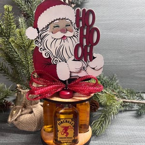 May include: A wooden Santa Claus figurine holding a sign that says "Ho Ho Oh" with a red bow and three mini bottles of Fireball Cinnamon Whisky.