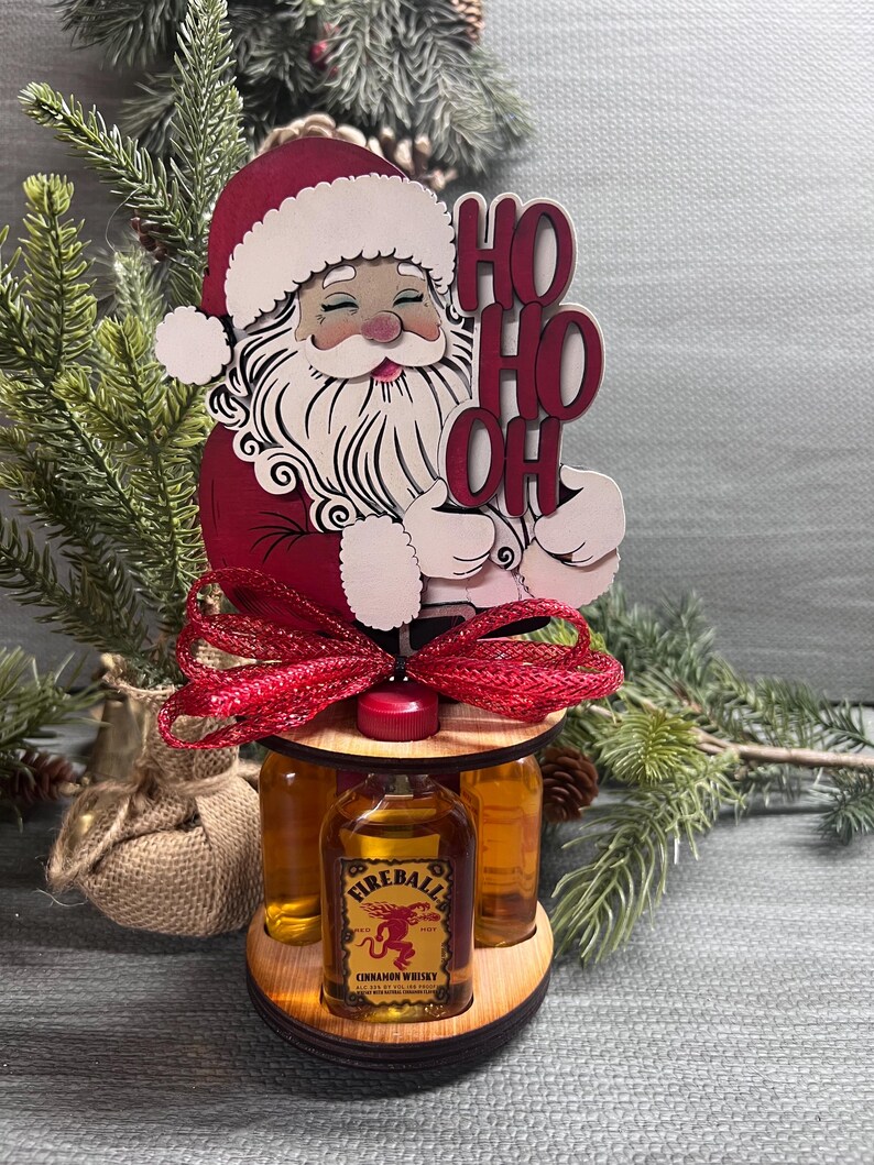 May include: A wooden Santa Claus figurine holding a sign that says "Ho Ho Oh" with a red bow and three mini bottles of Fireball Cinnamon Whisky.