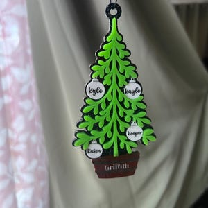 May include: A green Christmas tree ornament with black trim, featuring white ornaments with names: Kyle, Kayle, and Kooper. The tree sits in a brown pot with the name Griffith. The ornament hangs from a black loop.