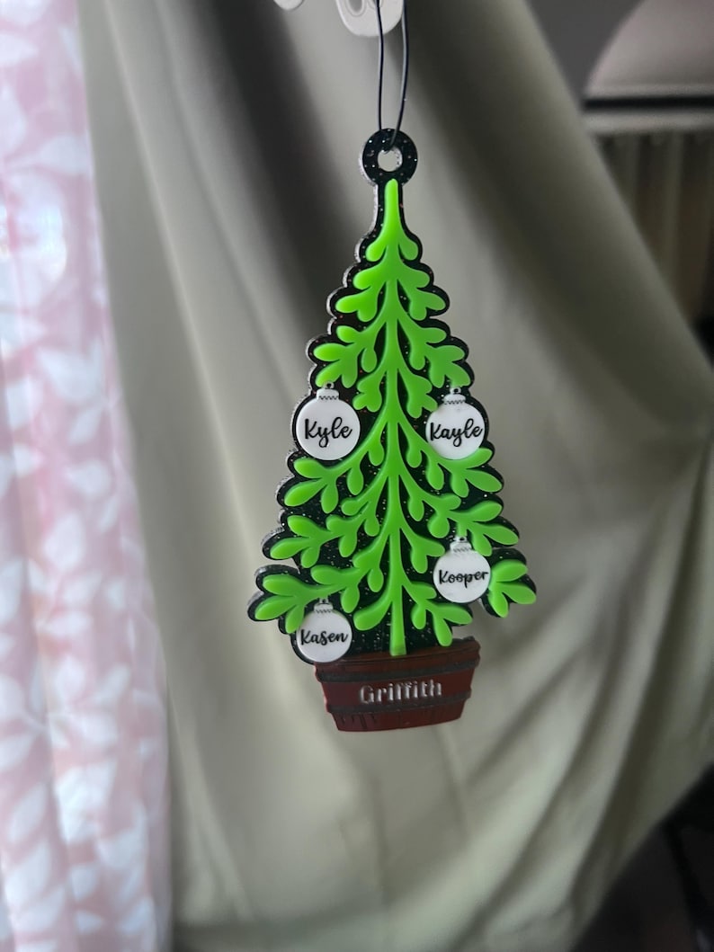 May include: A green Christmas tree ornament with black trim, featuring white ornaments with names: Kyle, Kayle, and Kooper. The tree sits in a brown pot with the name Griffith. The ornament hangs from a black loop.