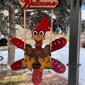 May include: A wooden sign featuring a cartoon rooster on a swing, with the word "Welcome" at the top. The rooster is red, brown, and yellow, with large eyes and outstretched wings. The sign is hanging outdoors.