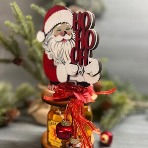 May include: A wooden Santa Claus figurine holding a sign that says "HO HO OH" is perched on top of a bottle of liquor. The bottle is decorated with a red ribbon and jingle bells.