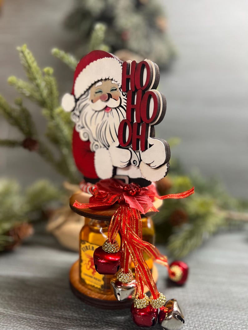May include: A wooden Santa Claus figurine holding a sign that says "HO HO OH" is perched on top of a bottle of liquor. The bottle is decorated with a red ribbon and jingle bells.