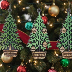 May include: Three wooden Christmas tree ornaments with green and black details, hanging on a decorated Christmas tree. Each ornament has a tree shape with names on round ornaments and the text "Merry Christmas 2023".
