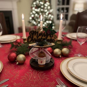 May include: A festive Christmas table setting with a red tablecloth and gold accents. A centerpiece features a bottle of Pendleton whisky, a "Merry Christmas" sign, and candles. Red and gold ornaments add to the holiday decor.