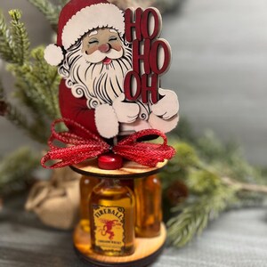 May include: Wooden Santa Claus figurine holding a sign that says "Ho Ho Oh" with a red bow and three mini bottles of Fireball Cinnamon Whisky.