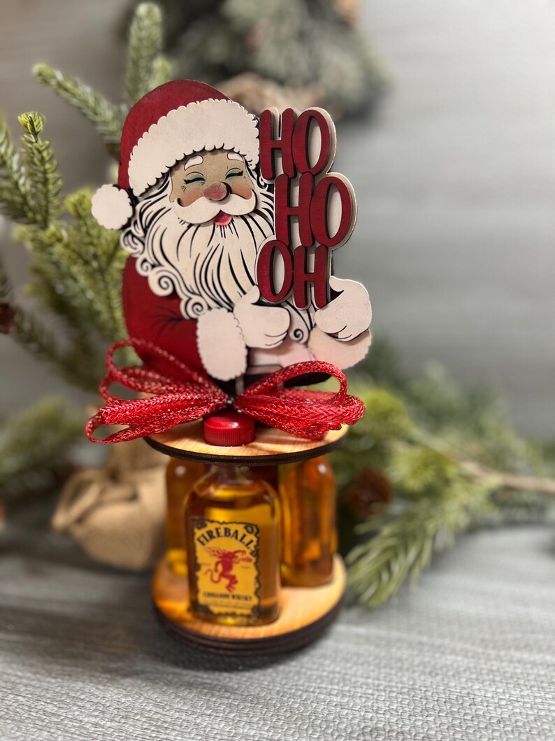 May include: Wooden Santa Claus figurine holding a sign that says "Ho Ho Oh" with a red bow and three mini bottles of Fireball Cinnamon Whisky.