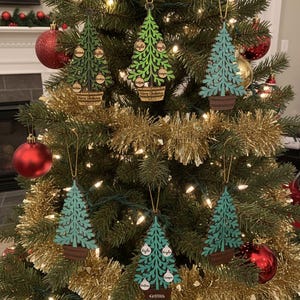 May include: Christmas tree ornaments in the shape of trees, hanging on a decorated Christmas tree. The ornaments are green and teal with family names on them. The ornaments are in a brown pot with the words "Merry Christmas" on them.