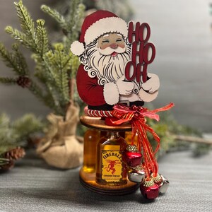 May include: A wooden Santa Claus figurine holding a sign that says "Ho Ho Oh" with two small bottles of Fireball Cinnamon Whisky on a wooden stand decorated with red ribbon and jingle bells.
