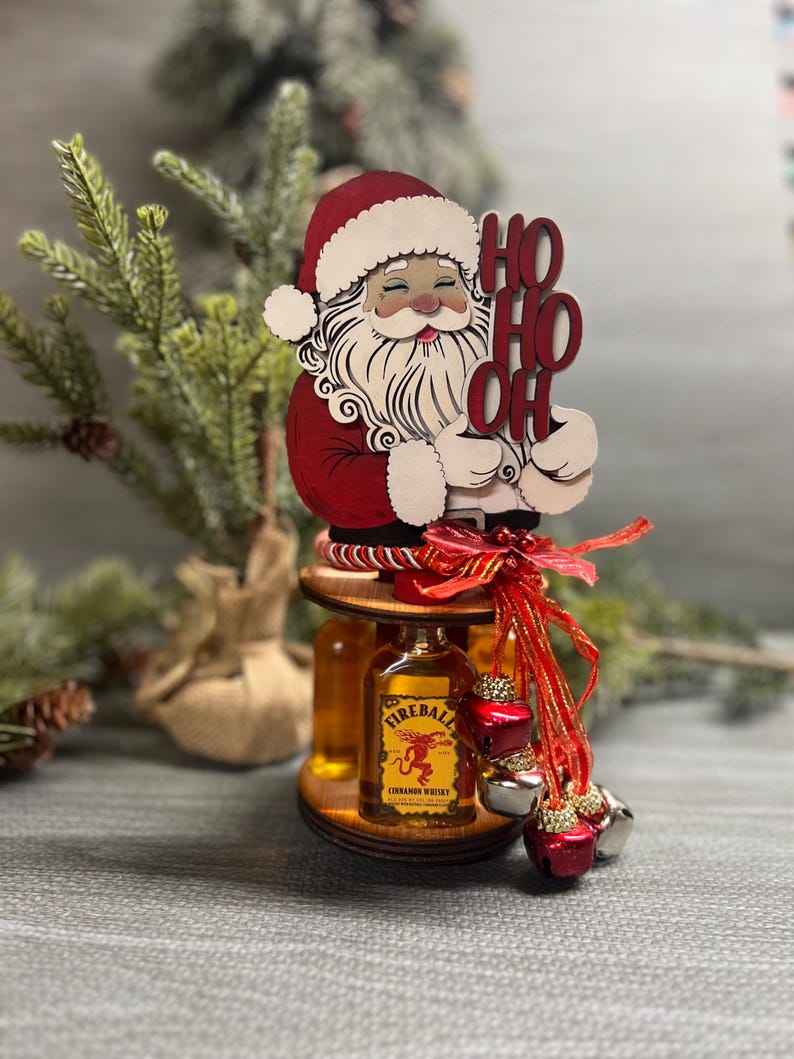 May include: A wooden Santa Claus figurine holding a sign that says "Ho Ho Oh" with two small bottles of Fireball Cinnamon Whisky on a wooden stand decorated with red ribbon and jingle bells.
