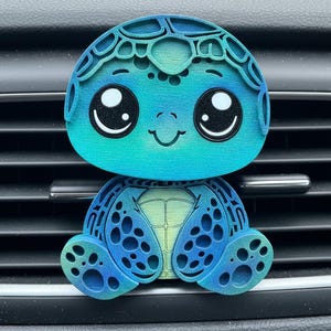 May include: A blue and green turtle-shaped car air vent clip. The turtle has large, expressive eyes, a smiling mouth, and a layered design. The color gradient shifts from light green to blue. The clip is designed to attach to a car's air vent.