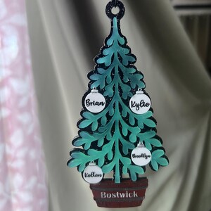 May include: A teal Christmas tree ornament with a black outline and glitter accents. The ornament has four white ball ornaments with names on them and a brown barrel base with the name "Bostwick".