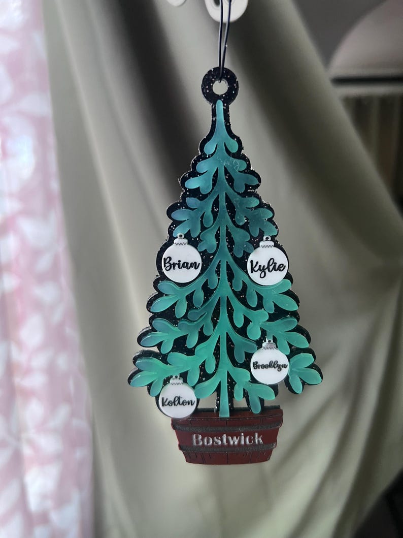 May include: A teal Christmas tree ornament with a black outline and glitter accents. The ornament has four white ball ornaments with names on them and a brown barrel base with the name "Bostwick".