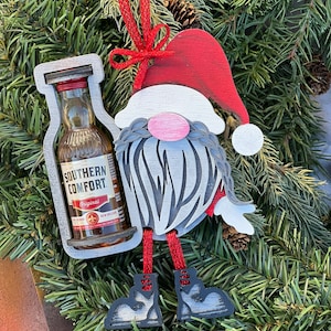 Puede incluir: Adorno navideño de madera con forma de gnomo que sostiene una botella en miniatura de licor Southern Comfort. El gnomo lleva un gorro rojo, barba blanca y botas rojas. La botella está etiquetada con "Southern Comfort Original."