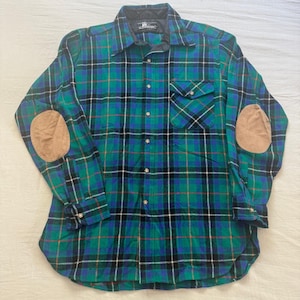 May include: A long-sleeve button-down shirt featuring a green, blue, and black plaid pattern. The shirt includes a single chest pocket, a dark collar, and tan-colored elbow patches on each sleeve. The front is secured with a series of buttons.