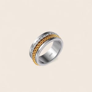Könnte beinhalten: Ein silberner Ring mit einem goldfarbenen Kettenband und einer Reihe klarer Steine. Der Ring hat ein rotierendes Design und ist mit "S925" markiert. Der Schmuck ist vor einem neutralen Hintergrund platziert.