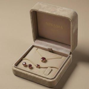 Puede incluir: Un conjunto de joyas de oro con piedras preciosas rojas, que incluye un collar, una pulsera y pendientes, presentado en una caja de terciopelo beige abierta. La caja tiene el nombre de la marca "NINASIA".