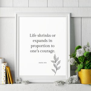 Op de afbeelding: Een witte ingelijste print met een zwart-wit citaat van Anaïs Nin: "Het leven krimpt of breidt zich uit in verhouding tot iemands moed."