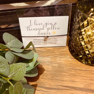 May include: A black bracelet with a small gold daisy charm. The bracelet has a message that reads "I love you a thousand yellow daisies. Close your eyes, make a wish, then put this bracelet on your wrist. When your bracelet breaks in two, your wish is ready to come true."