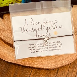 May include: A black string bracelet with a silver daisy charm. The bracelet is packaged with a white card that reads "I love you a thousand yellow daisies" and instructions for making a wish.