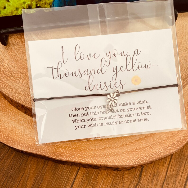 May include: A black string bracelet with a silver daisy charm. The bracelet is packaged with a white card that reads "I love you a thousand yellow daisies" and instructions for making a wish.